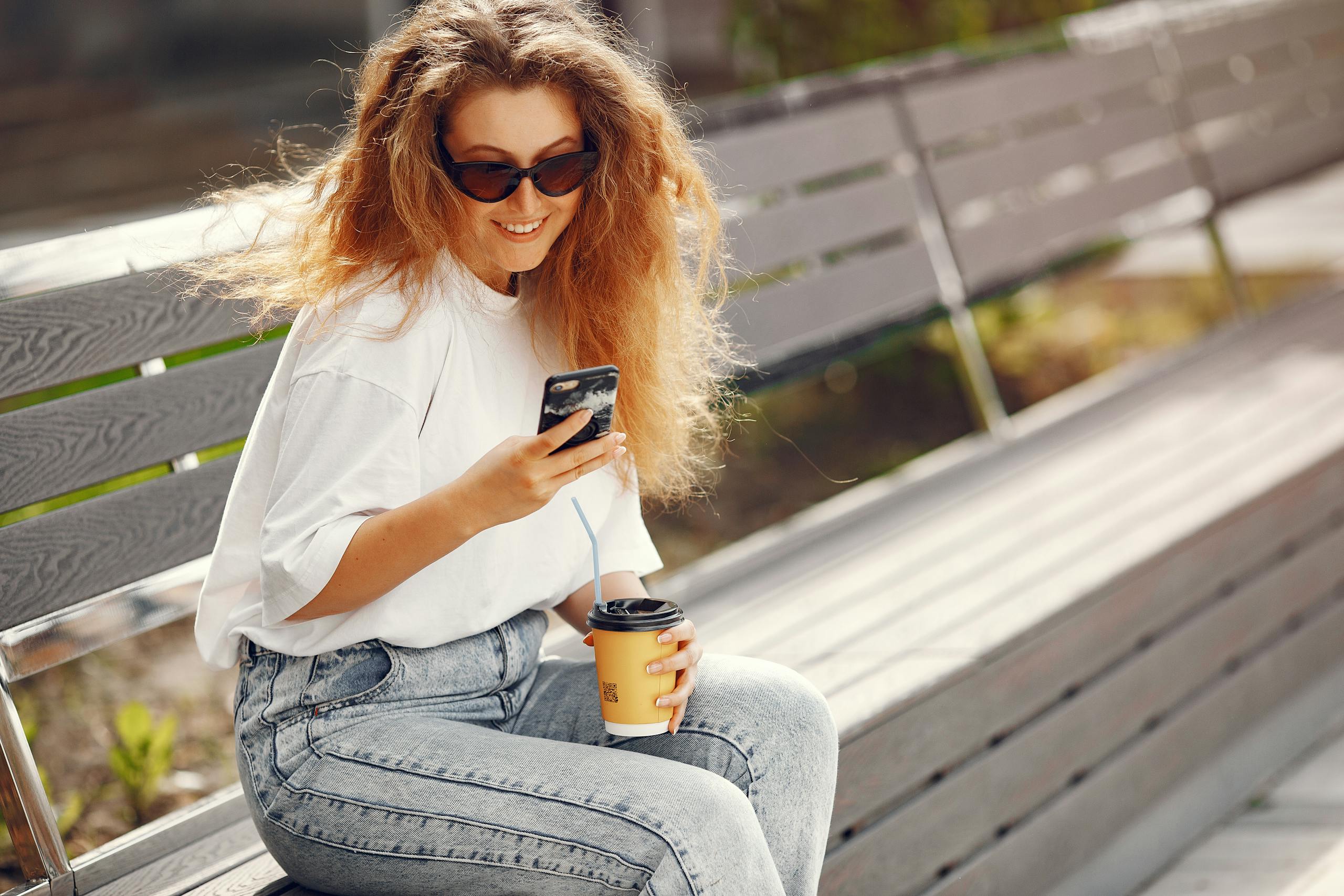 Casual young woman enjoying coffee while texting on a sunny park bench.